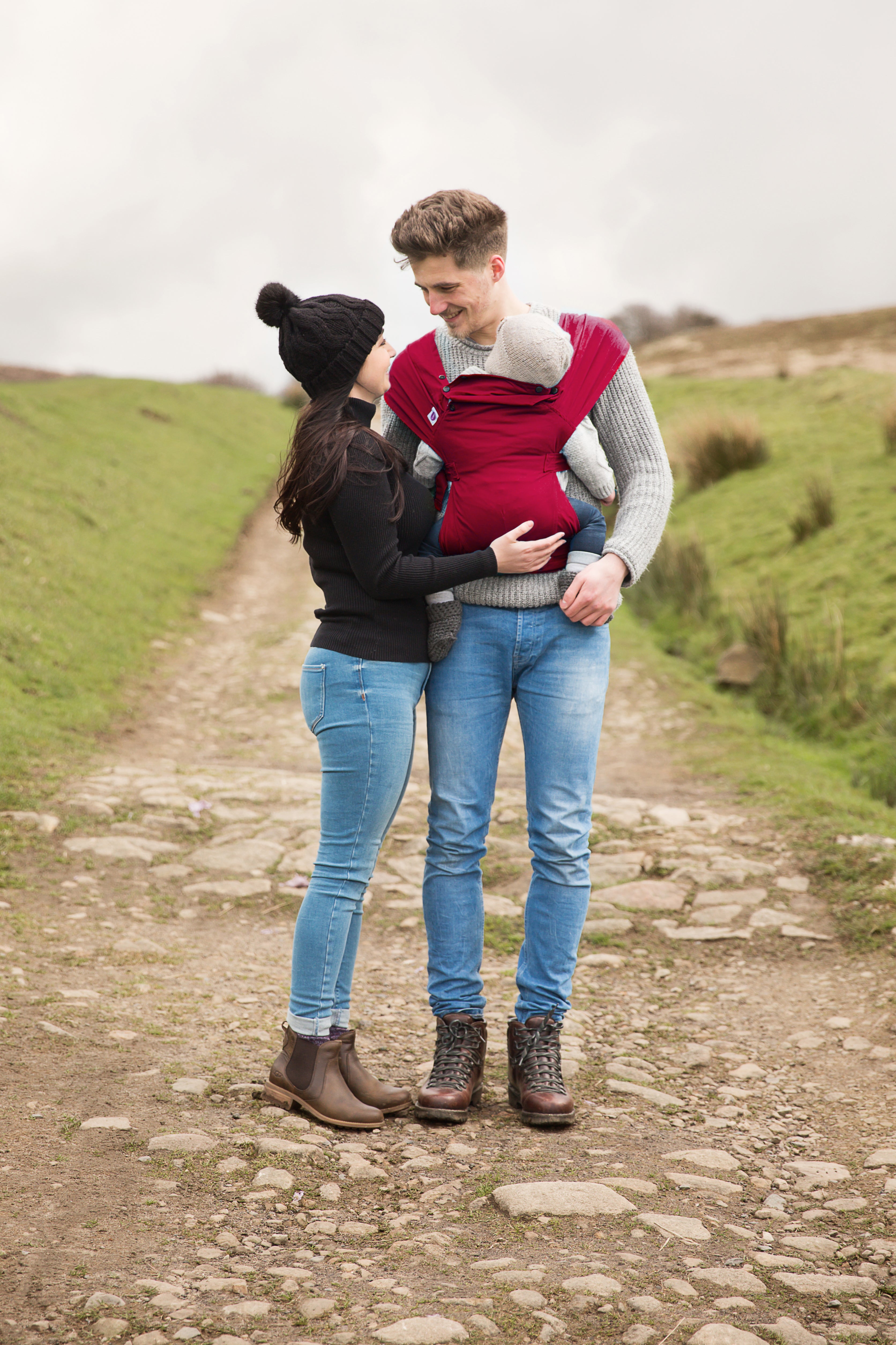 Ein Mann und eine Frau stehen gemeinsam auf einem steinigen Weg im Freien. Der Mann trägt ein Baby in der Babytrage Essential, die ergonomischen Halt bietet. Alle drei tragen Freizeitkleidung und scheinen glücklich in der Natur zu sein.