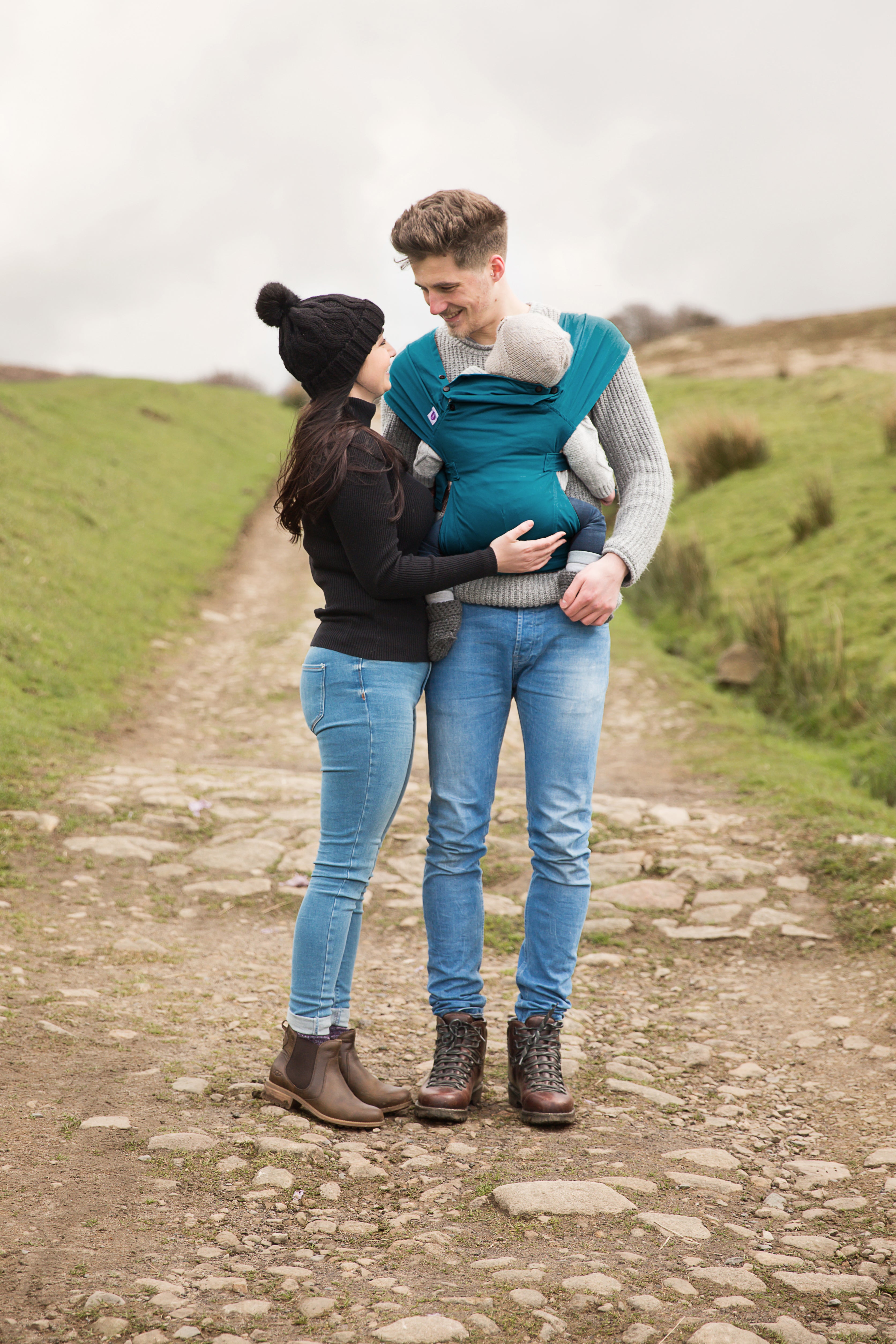 Ein Paar steht draußen auf einem steinigen Pfad. Die Frau in schwarzem Pullover und Mütze steht einem lächelnden Mann gegenüber, der ihr Baby in der Babytrage Essential trägt, die ergonomischen Halt für optimale Hüftgesundheit bietet. Beide tragen Jeans und Stiefel.