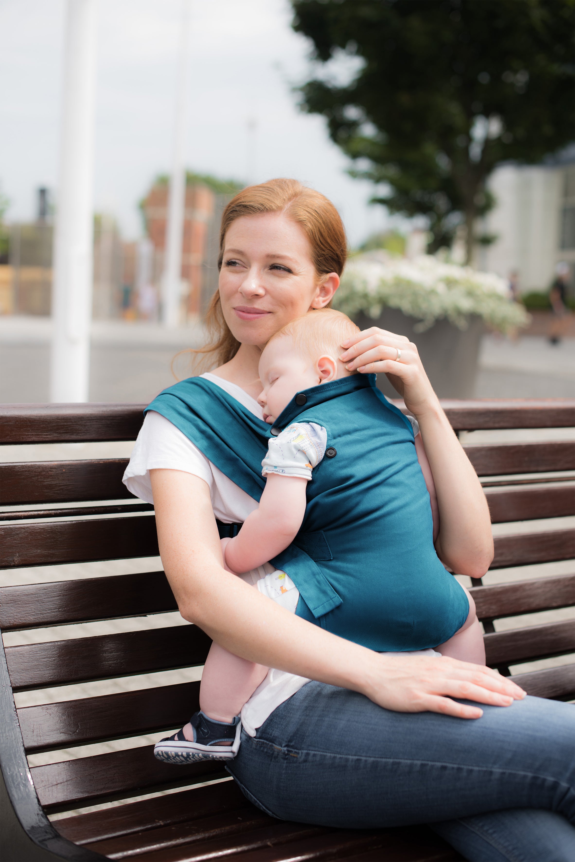 Eine Frau sitzt im Freien auf einer Holzbank und lächelt sanft, während sie ein schlafendes Baby in der blauen Babytrage Essential hält, die ergonomischen Halt bietet. Die Szene ist hell und friedlich.