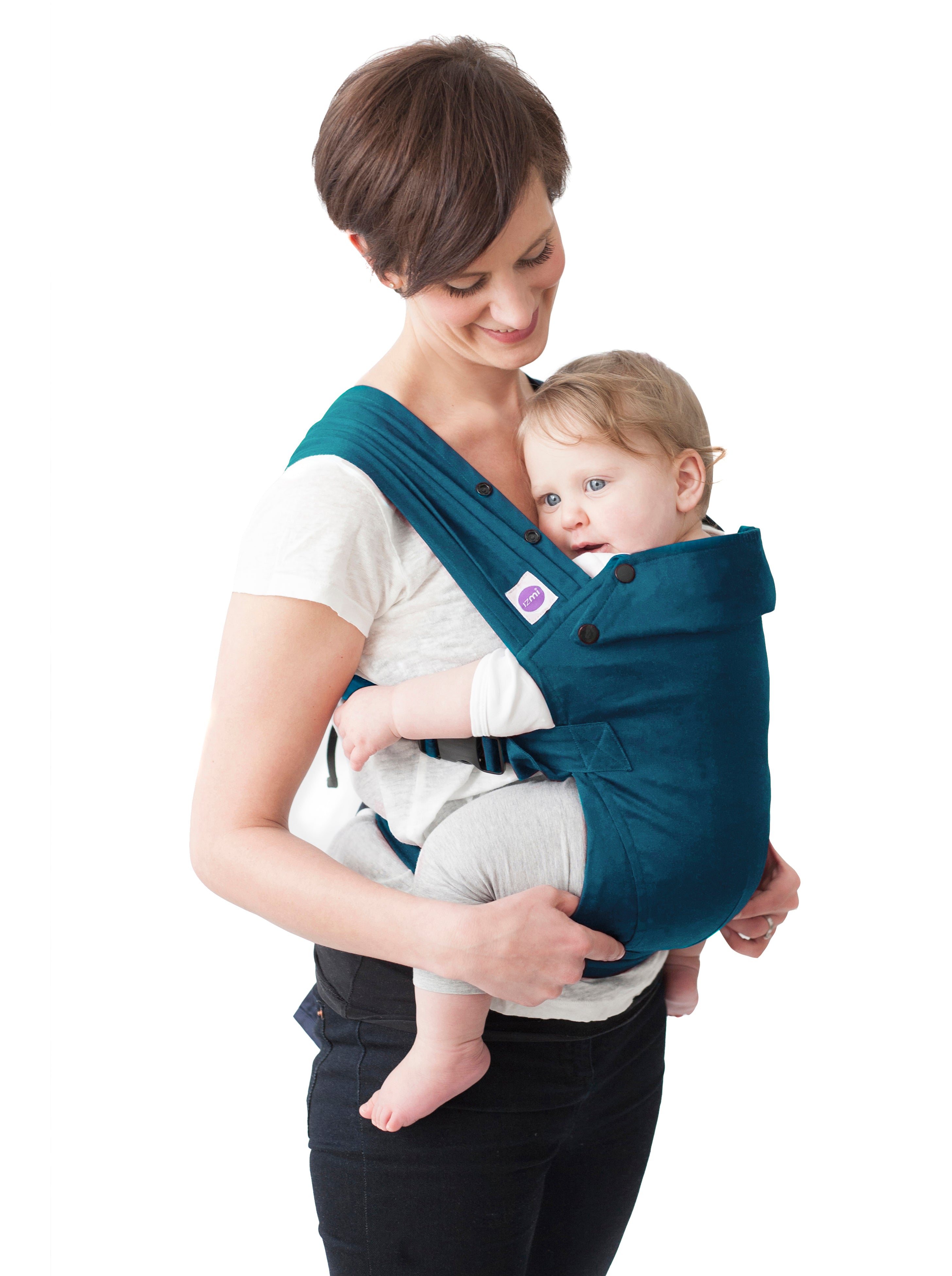 Eine Frau lächelt, während sie ihr Baby in der Babytrage Essential trägt, die ergonomische Unterstützung bietet und die Hüftgesundheit fördert. Das entspannte Baby blickt nach innen und trägt ein weißes Hemd und eine helle Hose vor einem weißen Hintergrund.