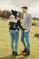 Eine Frau trägt ihr Baby in der Babytrage Breeze Grey mit Klimapaneel und hält dabei die Hand eines lächelnden Mannes. Beide tragen Pullover und Jeans draußen auf einer Wiese mit Bäumen, was den Komfort der Trage an einem kühlen Tag unterstreicht.