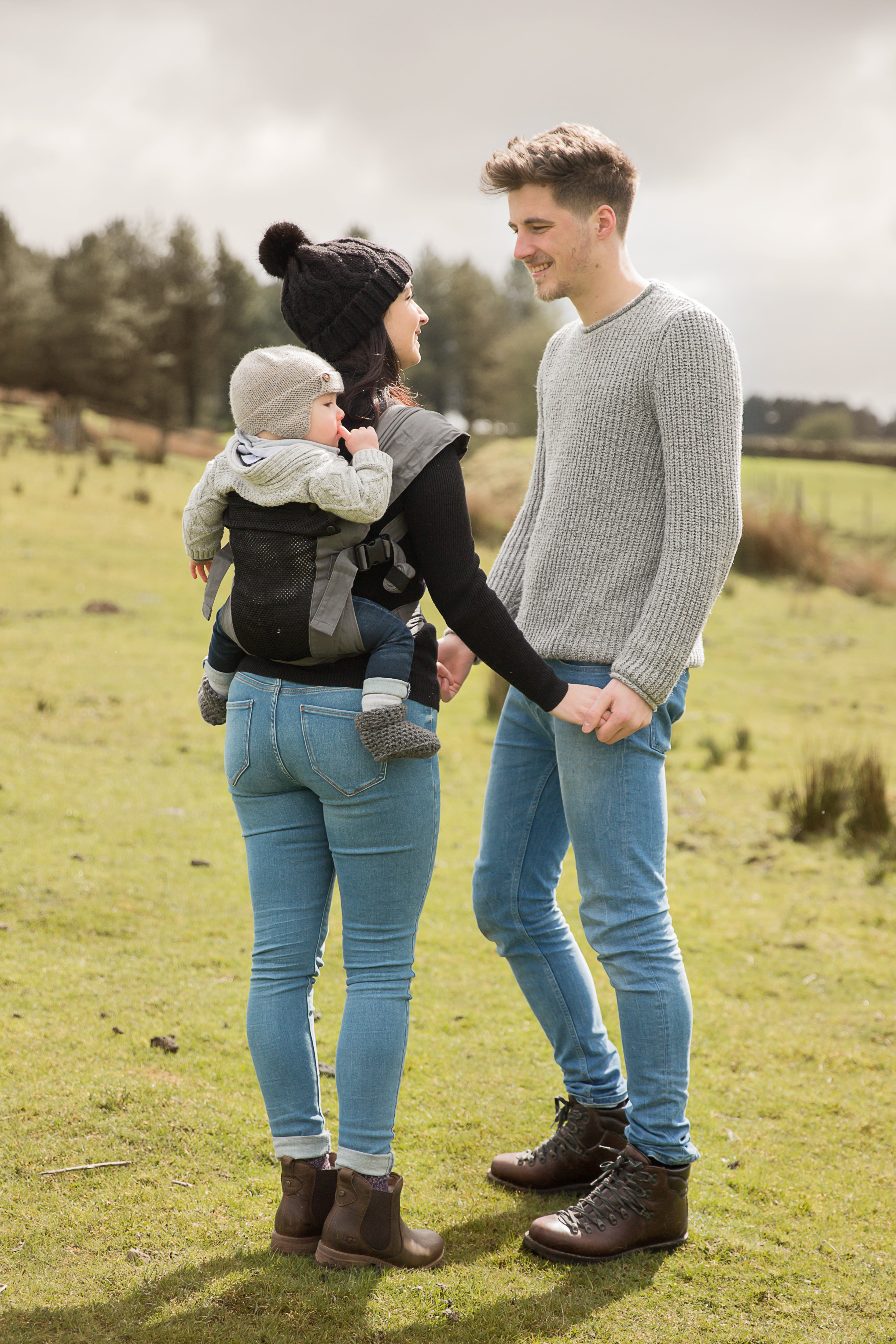 Eine Frau trägt ihr Baby in der Babytrage Breeze Grey mit Klimapaneel und hält dabei die Hand eines lächelnden Mannes. Beide tragen Pullover und Jeans draußen auf einer Wiese mit Bäumen, was den Komfort der Trage an einem kühlen Tag unterstreicht.
