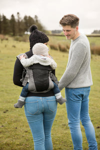 Eine Frau trägt ein Baby in der Babytrage Breeze Grey mit Klimapaneel, während ein leger gekleideter Mann daneben steht. Sie befinden sich an einem bewölkten Tag im Freien auf einem grasbewachsenen, offenen Feld.