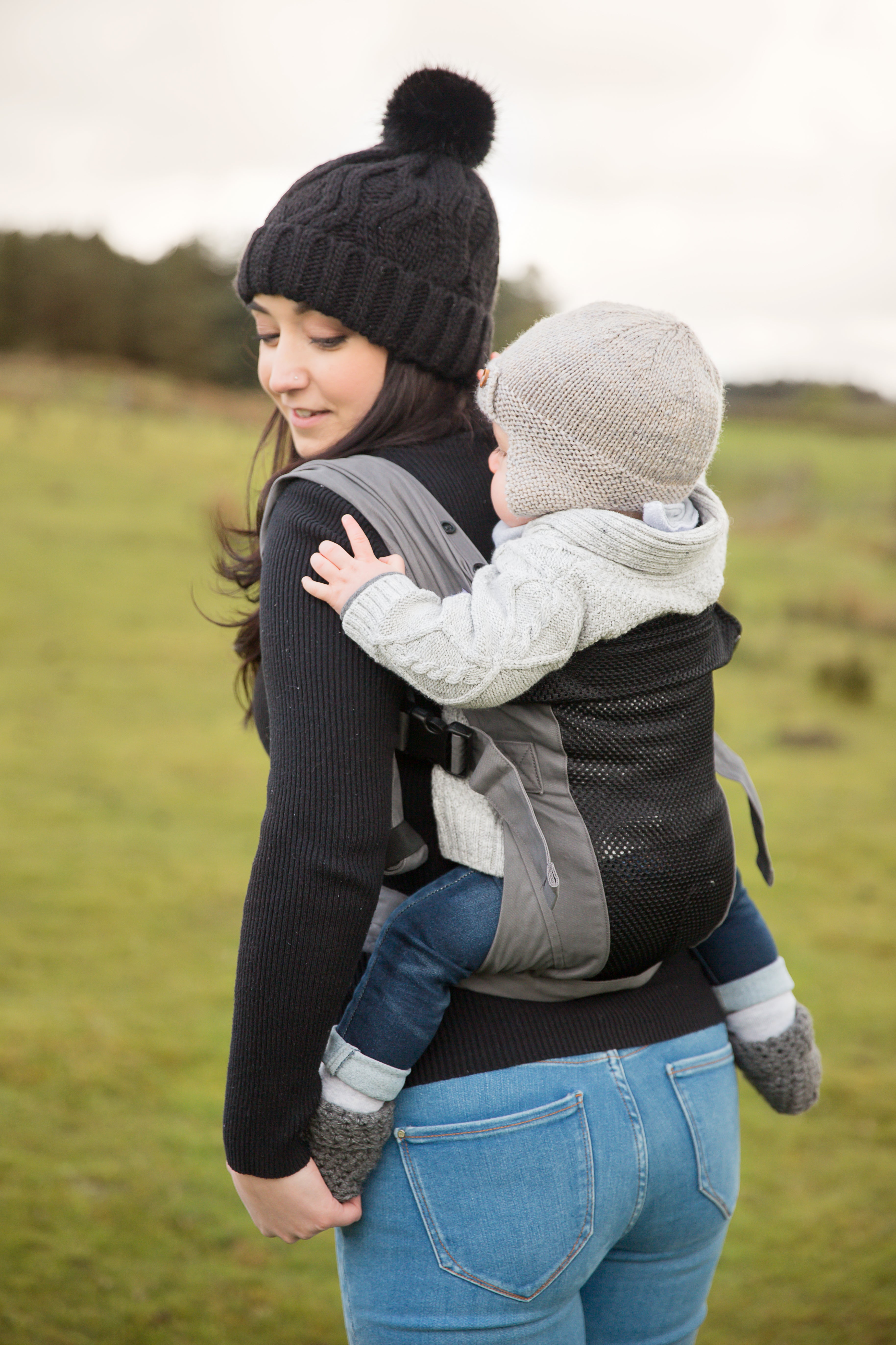Eine Frau mit einer schwarzen Strickmütze trägt ihr Baby, eingepackt in eine hellgraue Mütze und einen Pullover, in der Babytrage Breeze Grey mit Klimapaneel auf dem Rücken, während sie draußen auf einer Wiese die frische Luft genießen.