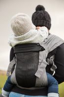 Ein Erwachsener mit dunkler Strickmütze und Pullover trägt ein Kind, beide warm angezogen, in einer Babytrage Breeze Grey mit Klimapaneel. Das Kind sitzt sicher mit den Armen um den Erwachsenen geschlungen und genießt den atmungsaktiven Komfort des Klimapaneels.
