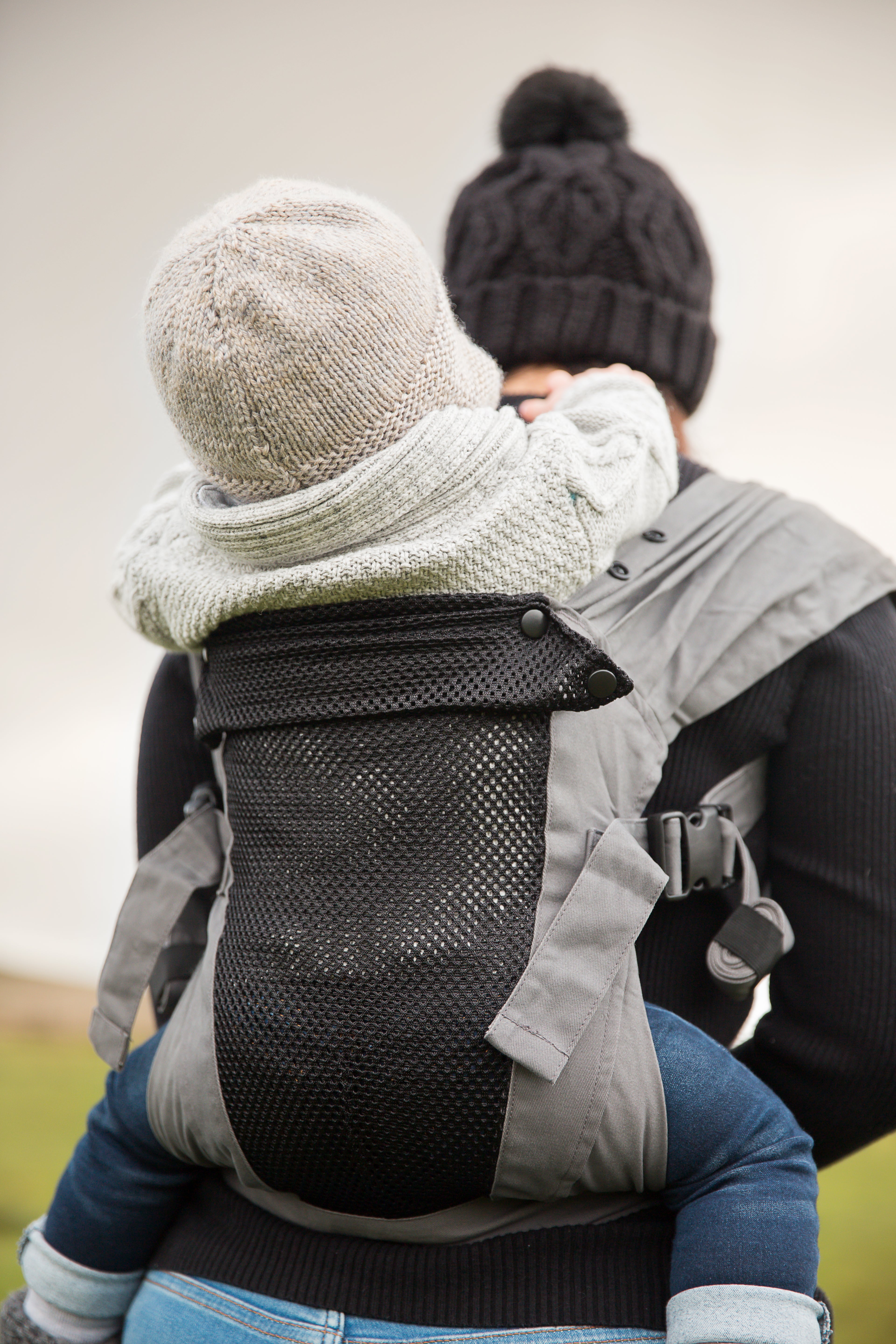 Ein Erwachsener mit dunkler Strickmütze und Pullover trägt ein Kind, beide warm angezogen, in einer Babytrage Breeze Grey mit Klimapaneel. Das Kind sitzt sicher mit den Armen um den Erwachsenen geschlungen und genießt den atmungsaktiven Komfort des Klimapaneels.