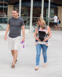 Ein Mann und eine Frau gehen vor einem Einkaufszentrum spazieren. Die Frau trägt ihr Baby in einer Babytrage Breeze Grey mit Klimapaneel. Der Mann hält eine Einkaufstasche in der Hand. Beide lächeln. Hinter ihnen sind Geschäfte und Glasgeländer zu sehen.
