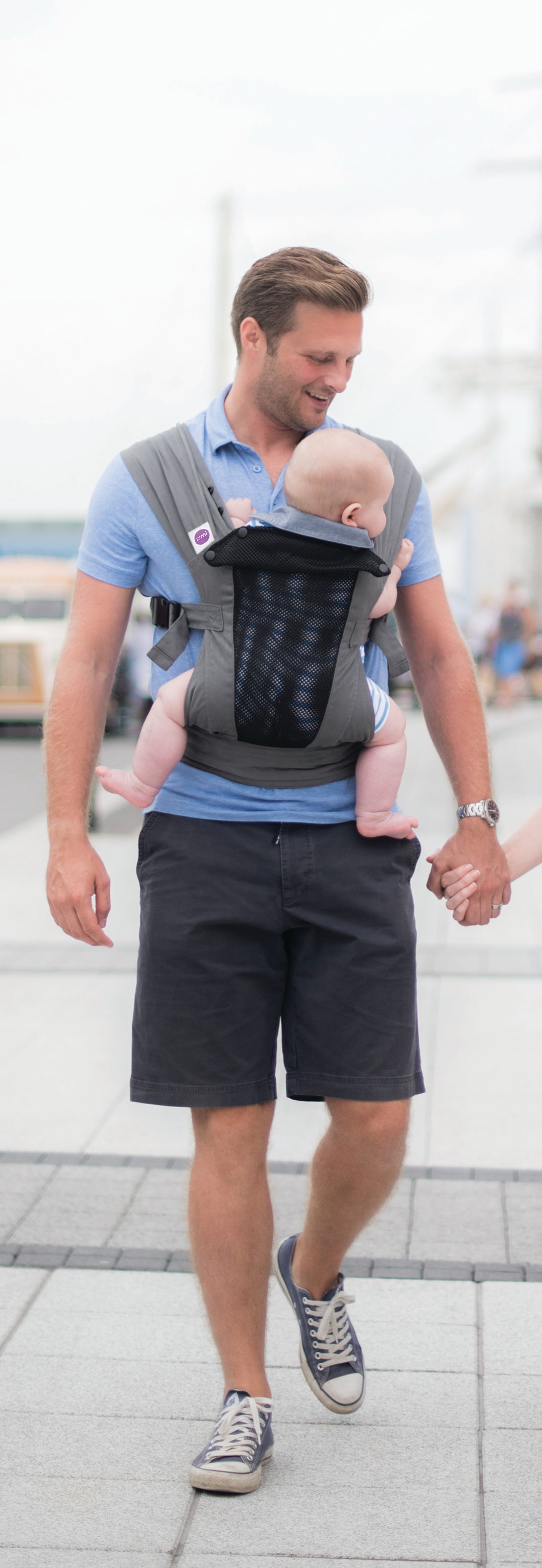 Ein Mann in blauem Hemd und dunklen Shorts geht im Freien spazieren. Er trägt ein Baby in einer Babytrage Breeze Grey mit Klimapaneel. Er lächelt das Baby an und hält die Hand einer anderen Person, deren Arm teilweise sichtbar ist.
