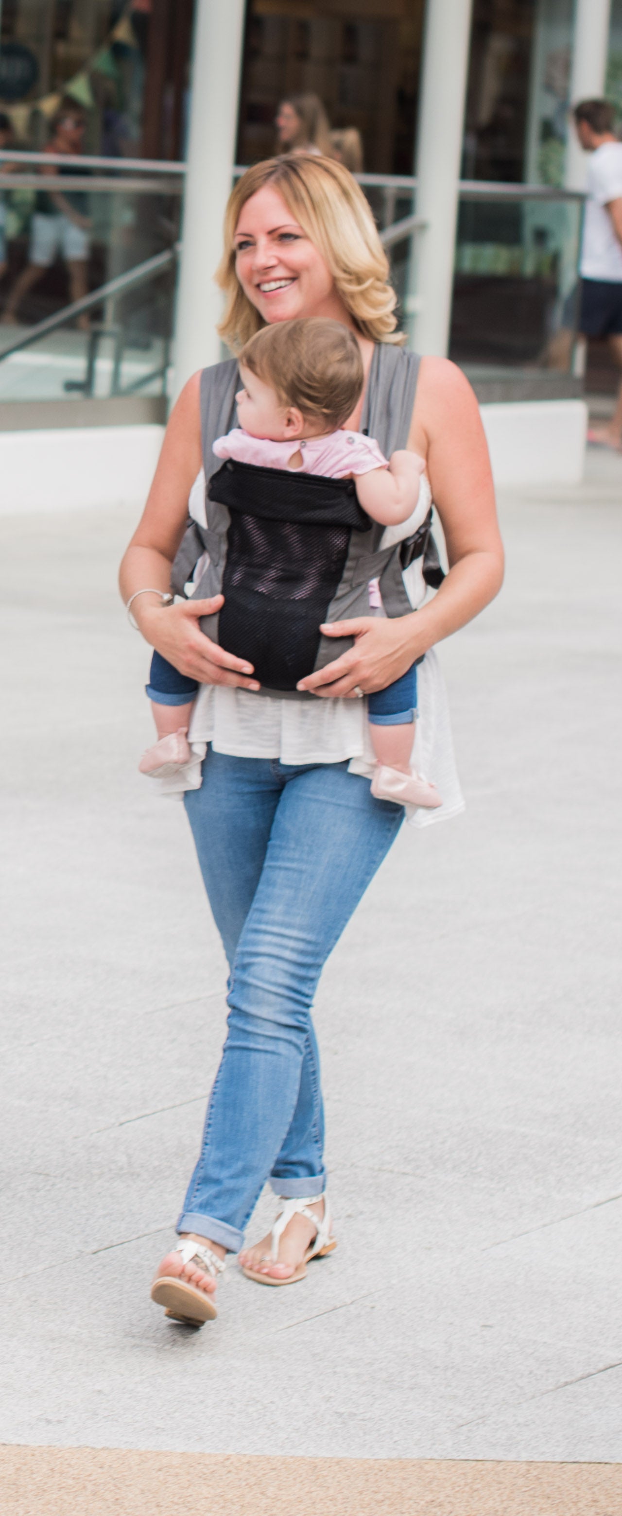 Eine lächelnde Frau trägt ihr Baby im Freien in der Babytrage Breeze Grey mit Klimapaneel, einem atmungsaktiven Mesh-Panel. Sie trägt ein ärmelloses Oberteil, Bluejeans und Sandalen; das Baby trägt helle Kleidung.