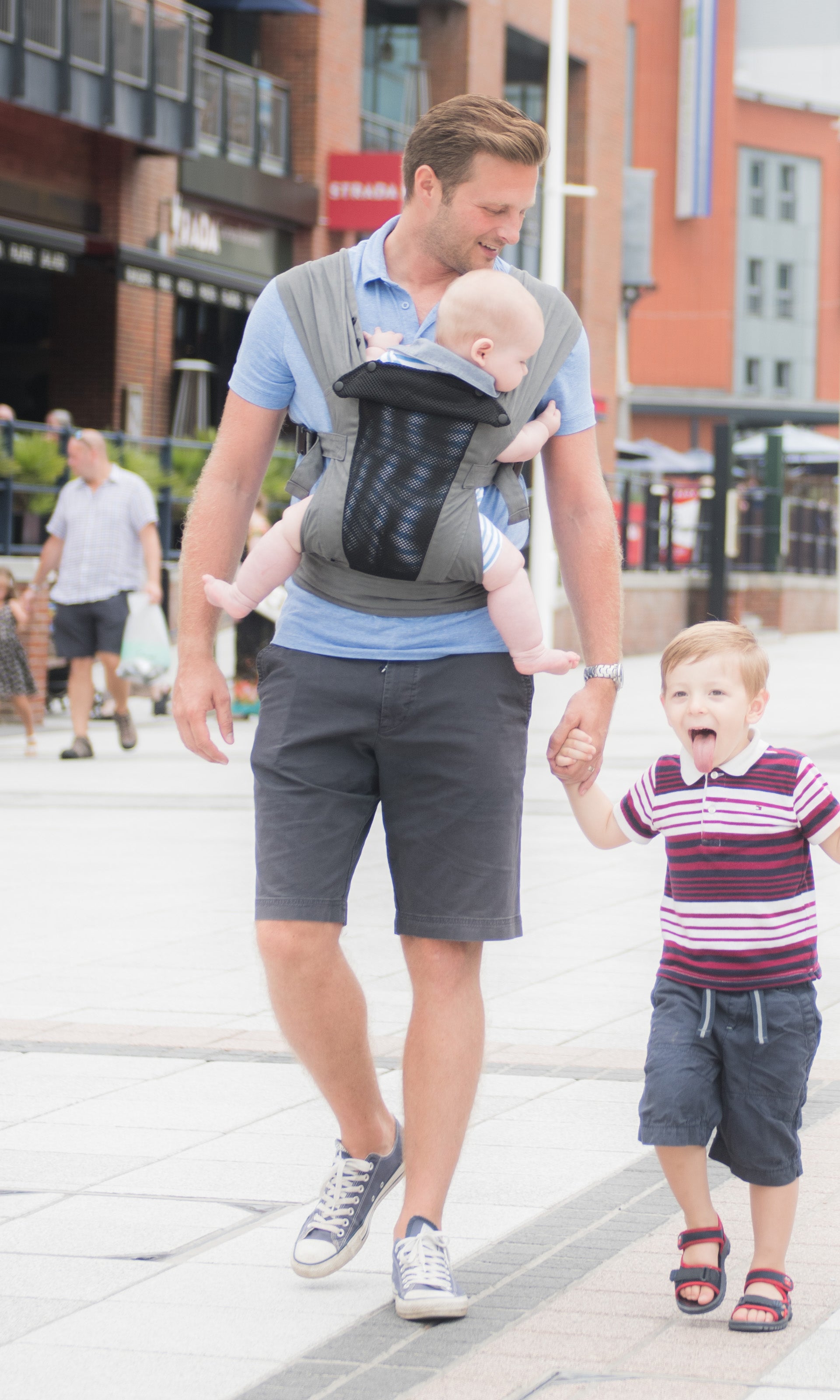 Ein Mann in Shorts und Turnschuhen geht draußen spazieren. Er trägt ein Baby in einer Babytrage Breeze Grey mit Klimapaneel und hält die Hand eines aufgeregten kleinen Jungen, der ihm die Zunge herausstreckt. Im Hintergrund sind weitere Menschen und Gebäude zu sehen.