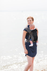 An einem sonnigen Strand lächelt eine Trageberaterin mit ihrem Baby in der Babytrage Breeze Grey mit Klimapaneel. Das atmungsaktive Mesh-Panel hält sie in ihrer leichten, lässigen Kleidung kühl und bequem.